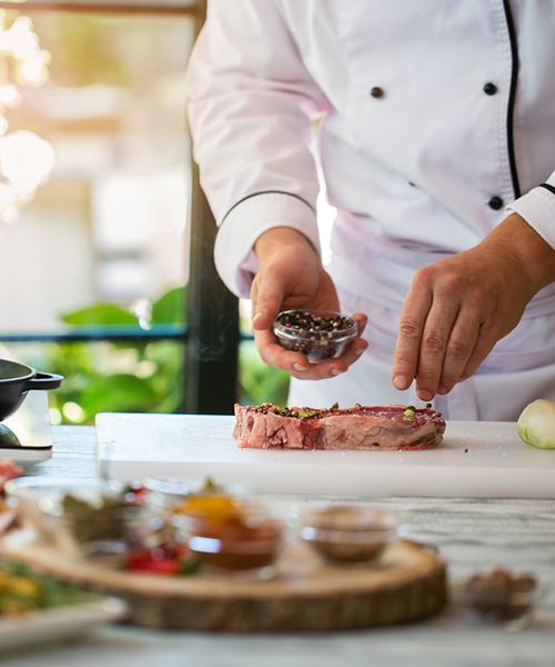 Chef sprinkling whole peppercorn on thick, juicy steak. Gourmet Spice Company, Wholesale Food Distributor.