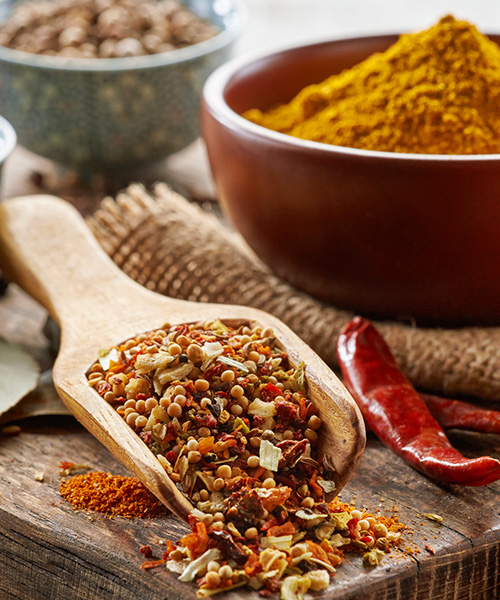 Dried spice blend in wooden spoon on thick cutting board with heaping bowl of turmeric in the background. Gourmet Spice Company, Wholesale Food Distributor.