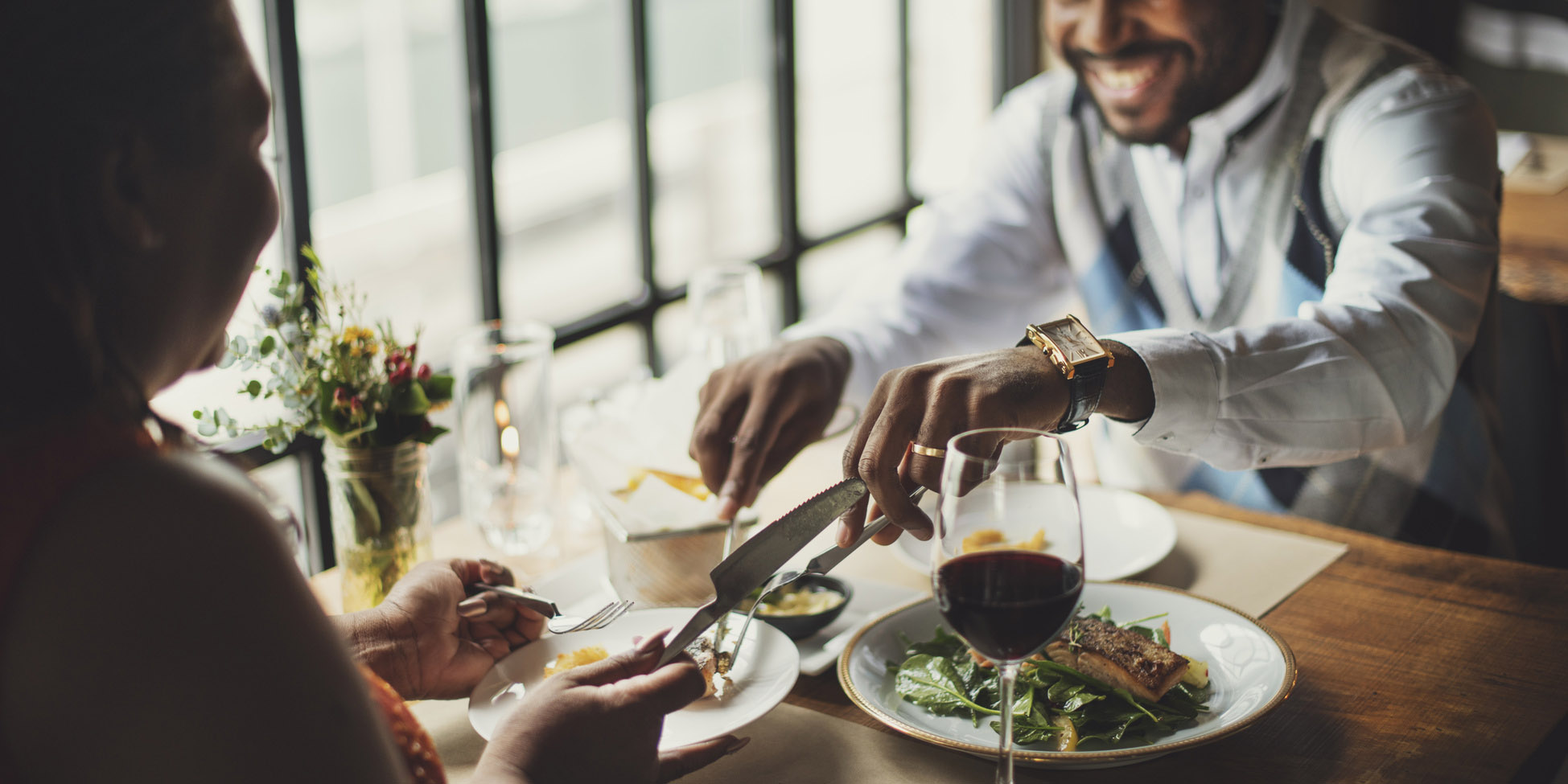 Couple smiling, enjoying red wine, and sharing plates of fish and fresh greens in hip restaurant. Gourmet Spice Company, Wholesale Food Distributor.