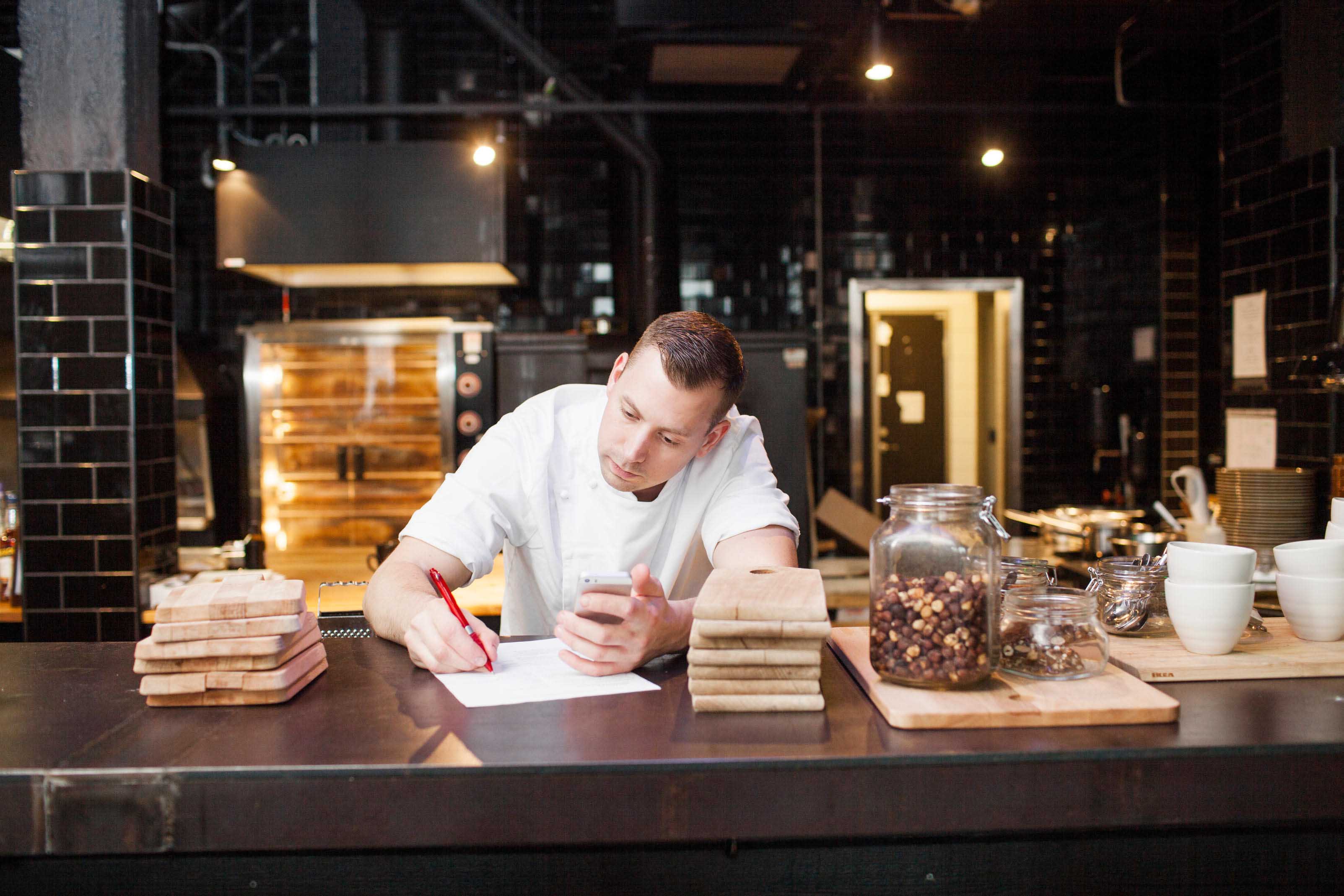 Chef in modern restaurant checking inventory list and ordering ingredients on his cell phone. Gourmet Spice Company, Wholesale Food Distributor.