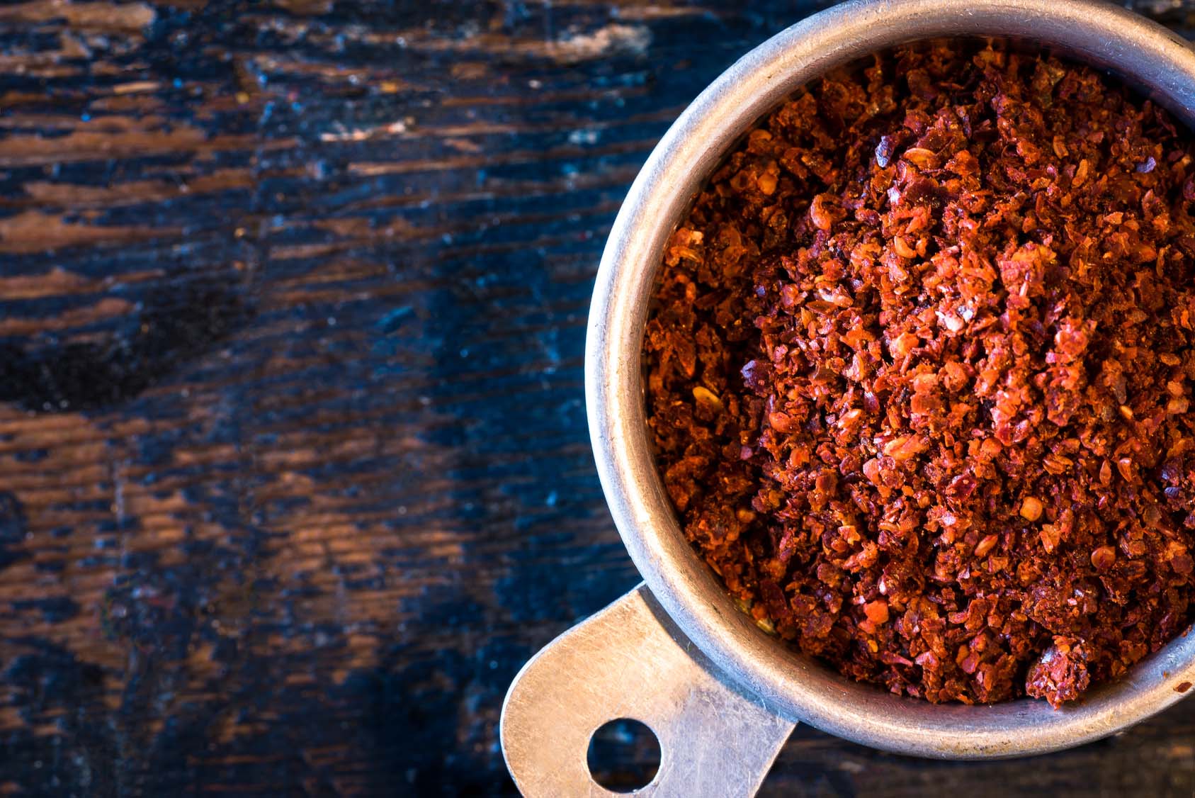 Bright red ground chili pepper in tin cup on wooden countertop. Gourmet Spice Company, Wholesale Food Distributor.