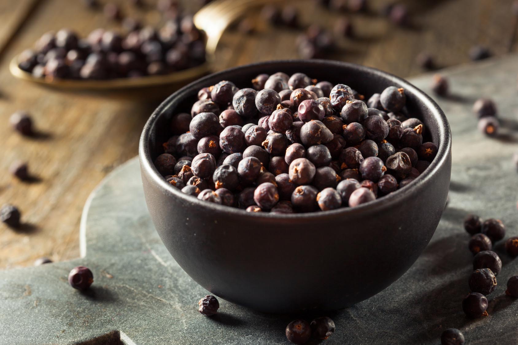 Dried juniper berries in bowl and scattered on granite cutting board. Gourmet Spice Company, Wholesale Food Distributor.