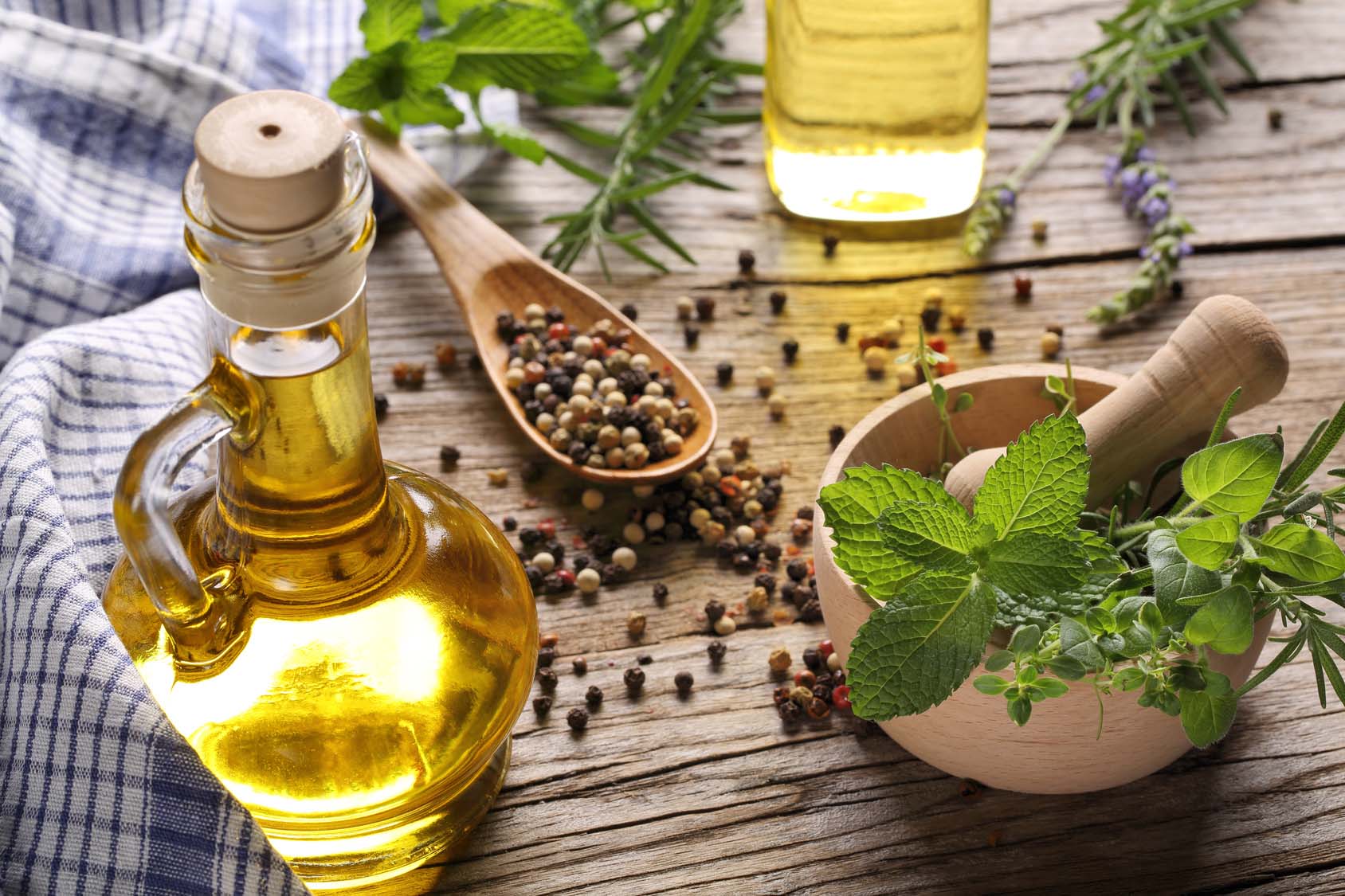 Extra virgin olive oil in glass jar next to peppercorns on wooden spoon, and fresh mint and herbs in a wooden mortar. Gourmet Spice Company, Wholesale Food Distributor.