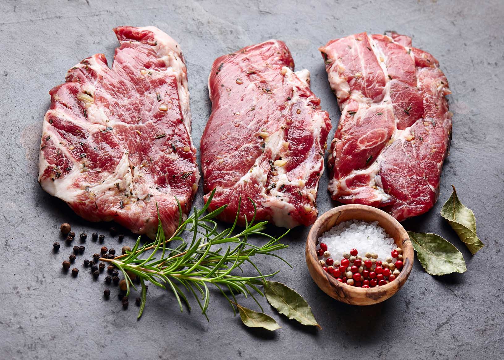 Three pork steaks on stone tabletop ready to cook, seasoned with fresh salt, peppercorn and rosemary. Gourmet Spice Company, Wholesale Food Distributor.